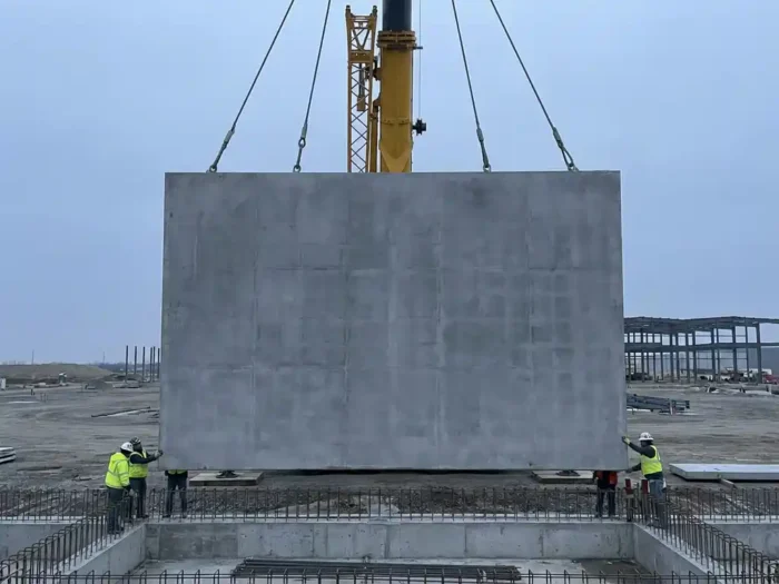 Grote betonnen wandplaat hangt aan industriële kraankabels tijdens plaatsing op bouwterrein onder bewolkte hemel.